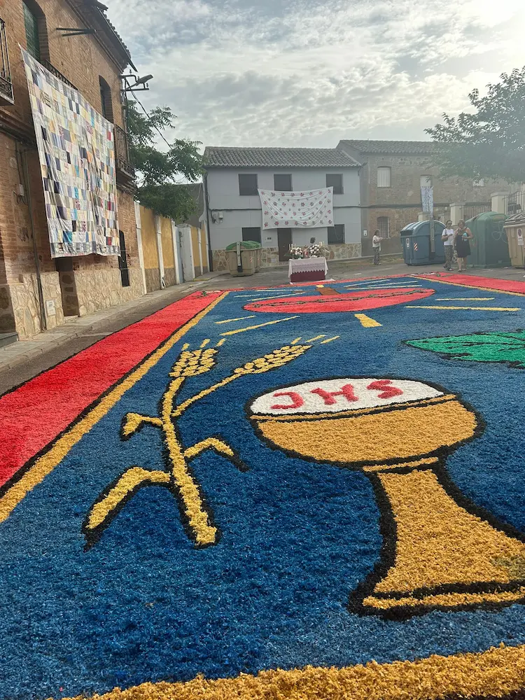Alfombra floral de Corpus Christi 2025 en Los Navalmorales con cáliz, espigas de trigo y formas litúrgicas en colores vivos sobre fondo azul