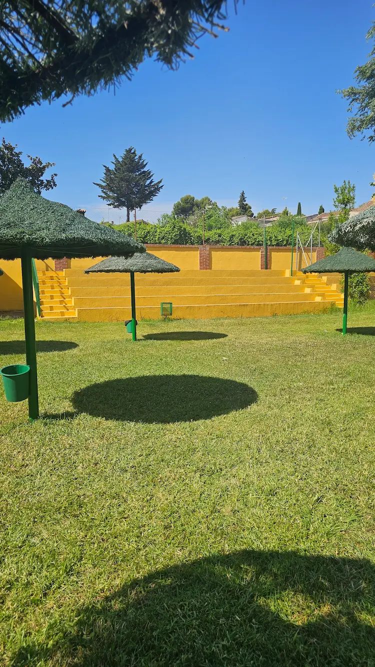 Zona verde con sombrillas y grada amarilla en la piscina municipal de Los Navalmorales, perfecta para relajarse al aire libre.